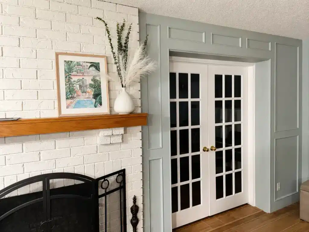 Painted white brick fireplace with natural wood mantel, decorative wall paneling, and white French doors in a modern Calgary living room.