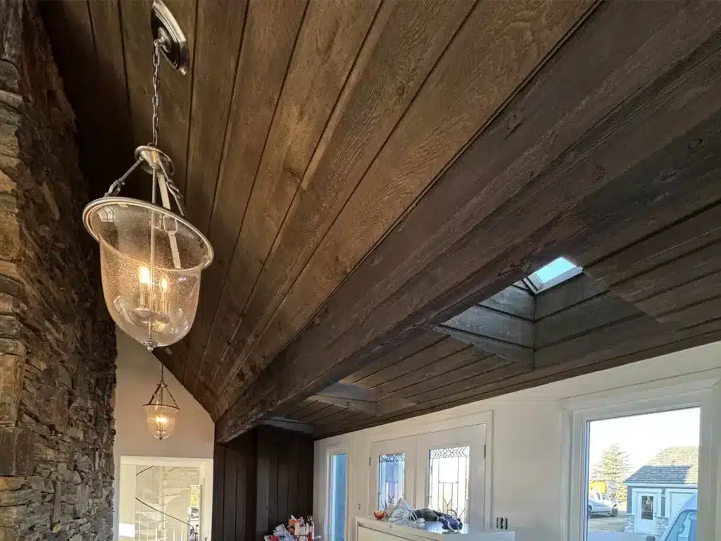 Dark stained wood plank ceiling with exposed beam, stone accent wall, skylight, and modern pendant lights in a custom home.